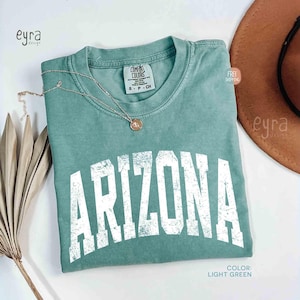 May include: A light green short-sleeve t-shirt with a crew neckline and the word ARIZONA printed in large, distressed white capital letters across the chest. The garment features a Comfort Colors brand label at the inner neck collar.