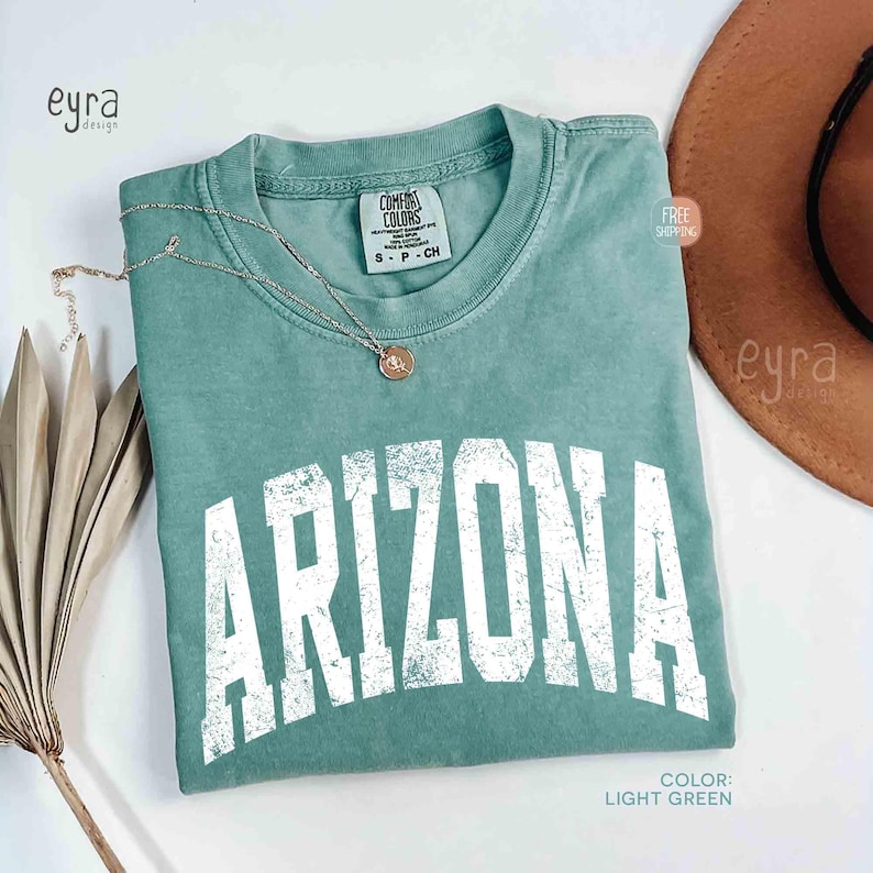 May include: A light green short-sleeve t-shirt with a crew neckline and the word ARIZONA printed in large, distressed white capital letters across the chest. The garment features a Comfort Colors brand label at the inner neck collar.