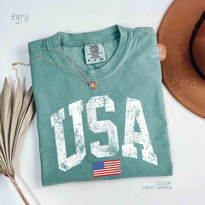 Puede incluir: Camiseta verde claro con la palabra "USA" en letras blancas desgastadas y una peque&ntilde;a bandera estadounidense. Un collar dorado con un colgante descansa sobre la camisa. La camisa est&aacute; doblada y se exhibe con un sombrero marr&oacute;n y una planta seca.