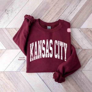 May include: A maroon-coloured sweatshirt featuring the text "KANSAS CITY" in distressed white lettering. The sweatshirt is folded on a light wood-grain surface. The colour is described as maroon.