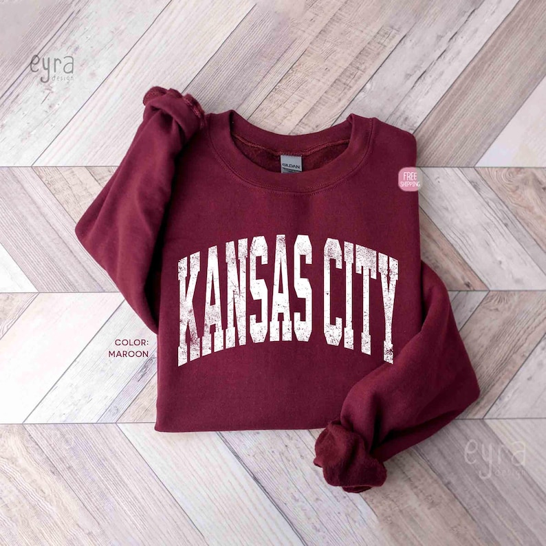 May include: A maroon-coloured sweatshirt featuring the text "KANSAS CITY" in distressed white lettering. The sweatshirt is folded on a light wood-grain surface. The colour is described as maroon.
