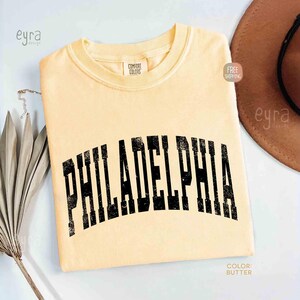 May include: A butter yellow short-sleeve t-shirt with a crew neckline. The shirt features the word PHILADELPHIA printed in a large, arched, black, distressed-style font across the chest. A Comfort Colors brand label is visible at the inner neck.