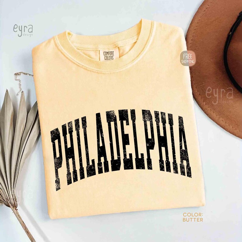 May include: A butter yellow short-sleeve t-shirt with a crew neckline. The shirt features the word PHILADELPHIA printed in a large, arched, black, distressed-style font across the chest. A Comfort Colors brand label is visible at the inner neck.