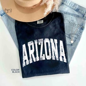 May include: A black short-sleeved t-shirt featuring the word ARIZONA printed in a large, distressed white collegiate-style font across the chest. The shirt has a crew neckline and a soft, washed-out appearance, showcasing a casual aesthetic.
