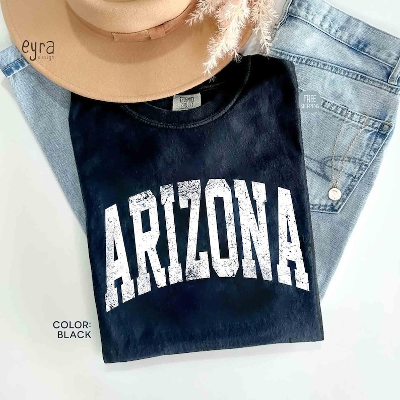 May include: A black short-sleeved t-shirt featuring the word ARIZONA printed in a large, distressed white collegiate-style font across the chest. The shirt has a crew neckline and a soft, washed-out appearance, showcasing a casual aesthetic.