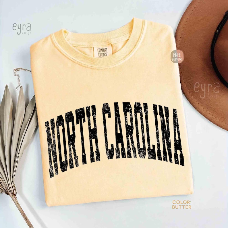 K&ouml;nnte beinhalten: Ein buttergelbes Kurzarm-T-Shirt mit Rundhalsausschnitt. Auf der Brust befindet sich der Schriftzug NORTH CAROLINA in einer gro&szlig;en, gebogenen und abgenutzten schwarzen Schriftart. Das Kleidungsst&uuml;ck hat ein Comfort Colors Etikett am Innenkragen.