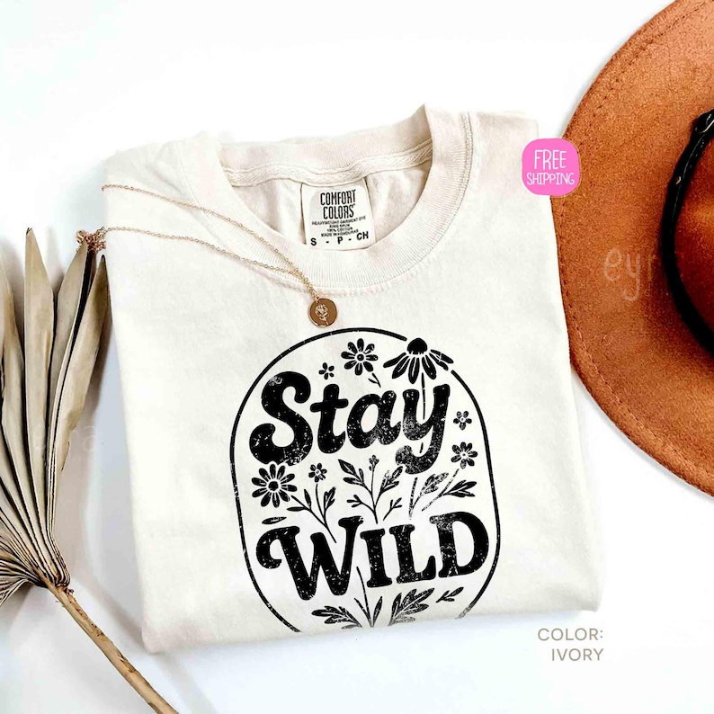 May include: Ivory-colored t-shirt with the words "Stay Wild" in a black floral design. The shirt is a Comfort Colors brand. A gold necklace and a brown hat are also in the image.
