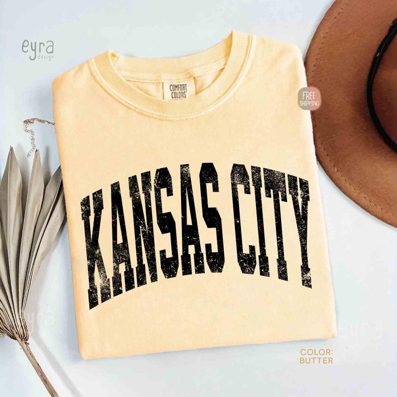May include: Butter-colored t-shirt with the text "KANSAS CITY" in distressed black lettering. The shirt is a Comfort Colors brand. The color of the shirt is butter.