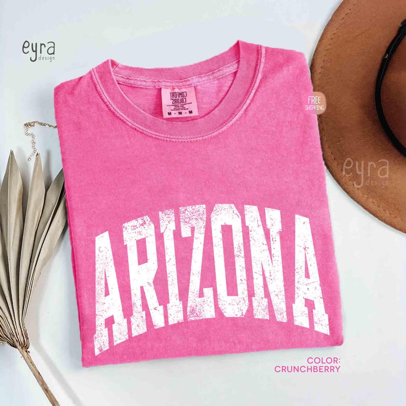 May include: A bright pink short-sleeve t-shirt with a crew neckline. The shirt features the word ARIZONA printed in large, distressed white capital letters across the chest. A tag inside the collar reads Comfort Colors with size M.