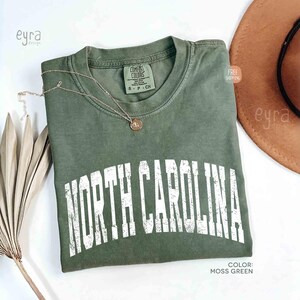 K&ouml;nnte beinhalten: Ein moosgr&uuml;nes Kurzarm-T-Shirt mit Rundhalsausschnitt und einem verwaschenen wei&szlig;en Grafikdruck, der in einer fetten, gebogenen Schrift NORTH CAROLINA zeigt. Das T-Shirt hat ein Markenetikett von Comfort Colors am Innenkragen und eine goldfarbene Kette.