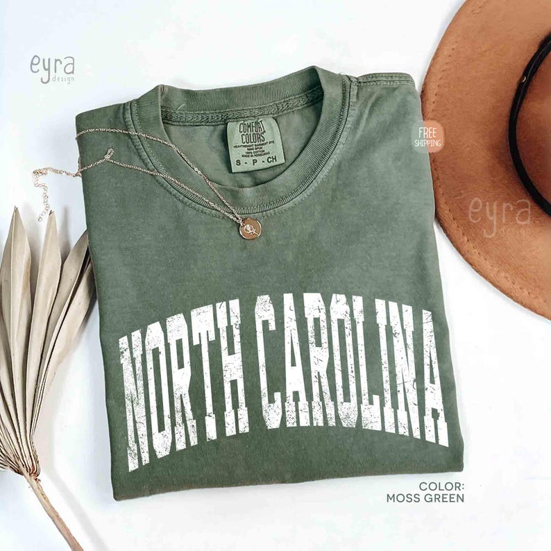 K&ouml;nnte beinhalten: Ein moosgr&uuml;nes Kurzarm-T-Shirt mit Rundhalsausschnitt und einem verwaschenen wei&szlig;en Grafikdruck, der in einer fetten, gebogenen Schrift NORTH CAROLINA zeigt. Das T-Shirt hat ein Markenetikett von Comfort Colors am Innenkragen und eine goldfarbene Kette.