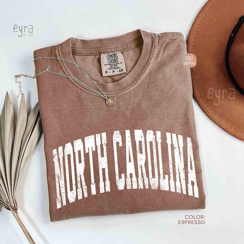 K&ouml;nnte beinhalten: Ein espressofarbenes Kurzarm-T-Shirt mit Rundhalsausschnitt und dem Schriftzug NORTH CAROLINA in verwaschener wei&szlig;er Schrift auf der Brust. Eine silberfarbene Halskette mit einem kleinen runden Anh&auml;nger liegt auf dem T-Shirt-Kragen.