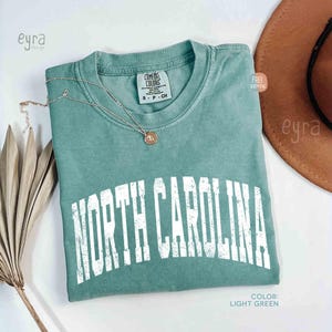 K&ouml;nnte beinhalten: Ein hellgr&uuml;nes Kurzarm-T-Shirt mit einem verwaschenen, wei&szlig;en, bogenf&ouml;rmigen Schriftzug NORTH CAROLINA. Das Shirt hat einen Rundhalsausschnitt und ein Comfort Colors Markenetikett am Innenkragen, kombiniert mit einer d&uuml;nnen goldfarbenen Halskette.