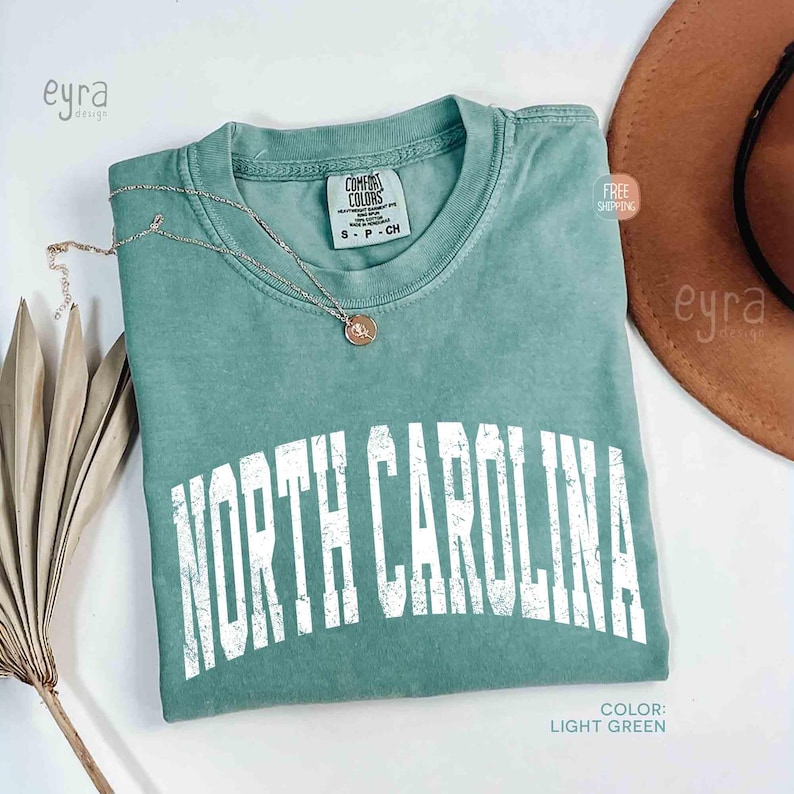 K&ouml;nnte beinhalten: Ein hellgr&uuml;nes Kurzarm-T-Shirt mit einem verwaschenen, wei&szlig;en, bogenf&ouml;rmigen Schriftzug NORTH CAROLINA. Das Shirt hat einen Rundhalsausschnitt und ein Comfort Colors Markenetikett am Innenkragen, kombiniert mit einer d&uuml;nnen goldfarbenen Halskette.