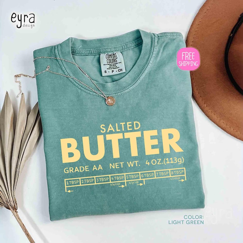 May include: Light green t-shirt with the text "SALTED BUTTER" in yellow. The shirt also features a graphic with measurements and the text "GRADE AA NET WT. 4 OZ. (113g)". A silver necklace is draped on the shirt.