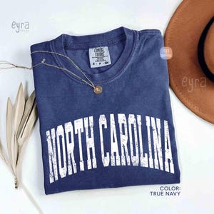 K&ouml;nnte beinhalten: Ein kurz&auml;rmeliges T-Shirt in Marineblau mit einem verwaschenen wei&szlig;en Grafikdruck, der NORTH CAROLINA in einer gro&szlig;en, gebogenen Schrift zeigt. Das Shirt hat ein Comfort Colors Markenetikett am Innenhalsausschnitt und einen Rundhalsausschnitt.