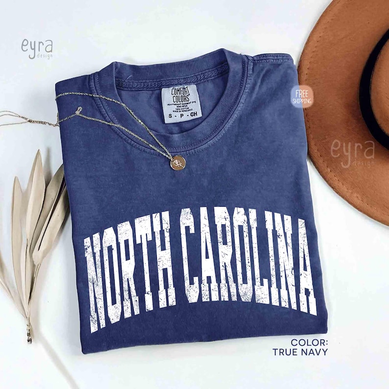 K&ouml;nnte beinhalten: Ein kurz&auml;rmeliges T-Shirt in Marineblau mit einem verwaschenen wei&szlig;en Grafikdruck, der NORTH CAROLINA in einer gro&szlig;en, gebogenen Schrift zeigt. Das Shirt hat ein Comfort Colors Markenetikett am Innenhalsausschnitt und einen Rundhalsausschnitt.