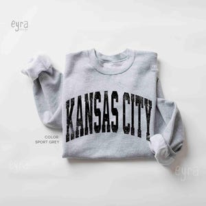 May include: A light grey sweatshirt featuring the text "KANSAS CITY" in distressed black lettering. The sweatshirt is sport grey in colour and has rolled-up sleeves.