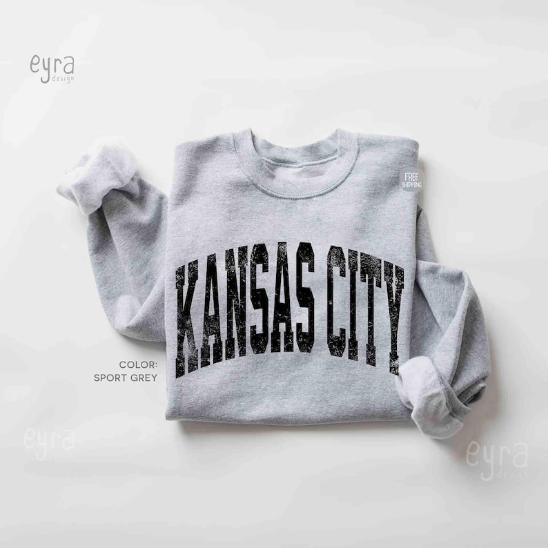 May include: A light grey sweatshirt featuring the text "KANSAS CITY" in distressed black lettering. The sweatshirt is sport grey in colour and has rolled-up sleeves.