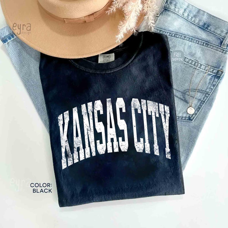 May include: Black t-shirt with the text "KANSAS CITY" in white, distressed lettering. The shirt is paired with light-wash denim jeans and a tan fedora hat. The color of the shirt is black.
