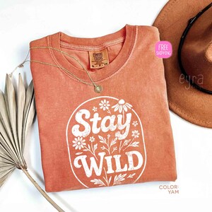 May include: A coral-colored t-shirt with the words "Stay Wild" in white, surrounded by floral designs. A gold necklace and a brown hat are also visible. The shirt has a crew neck and a Comfort Colors tag.