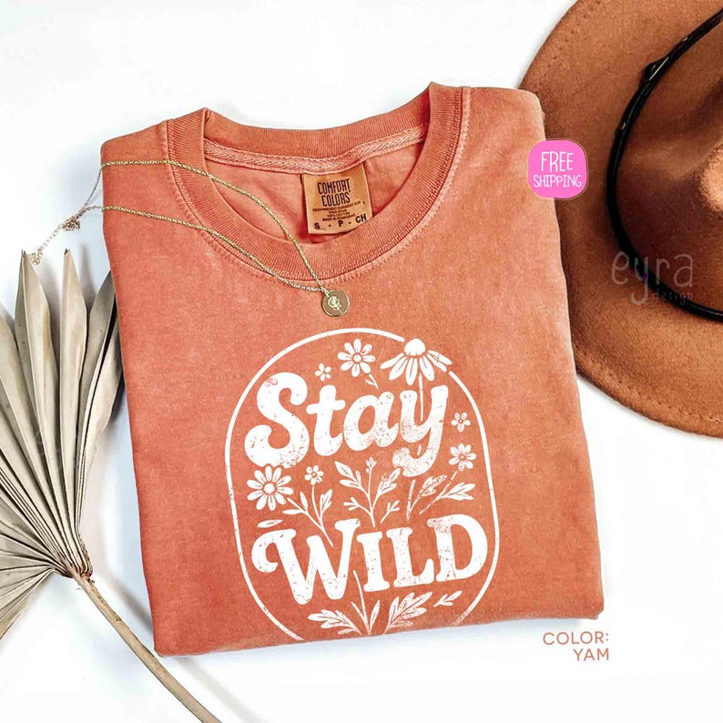 May include: A coral-colored t-shirt with the words "Stay Wild" in white, surrounded by floral designs. A gold necklace and a brown hat are also visible. The shirt has a crew neck and a Comfort Colors tag.