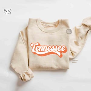 K&ouml;nnte beinhalten: Ein sandfarbenes Sweatshirt mit Rundhalsausschnitt und dem Wort Tennessee, das in einer kr&auml;ftigen, orangefarbenen Schrift im Retro-Stil auf der Brust gedruckt ist. Das Kleidungsst&uuml;ck hat lange &Auml;rmel sowie gerippte B&uuml;ndchen und einen Saum.