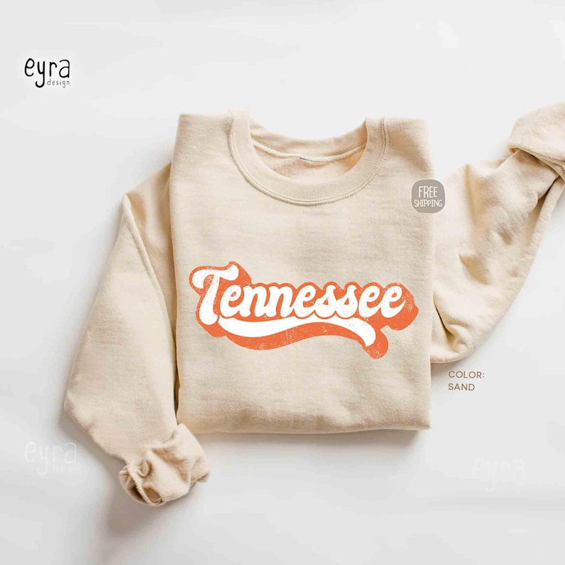 K&ouml;nnte beinhalten: Ein sandfarbenes Sweatshirt mit Rundhalsausschnitt und dem Wort Tennessee, das in einer kr&auml;ftigen, orangefarbenen Schrift im Retro-Stil auf der Brust gedruckt ist. Das Kleidungsst&uuml;ck hat lange &Auml;rmel sowie gerippte B&uuml;ndchen und einen Saum.