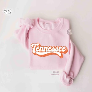 K&ouml;nnte beinhalten: Ein hellrosa Rundhals-Sweatshirt mit dem Wort Tennessee, das in einer kr&auml;ftigen, orangefarbenen Retro-Schrift mit wei&szlig;er Umrandung aufgedruckt ist. Das Kleidungsst&uuml;ck hat lange &Auml;rmel und einen gerippten Kragen mit einem klaren Design.