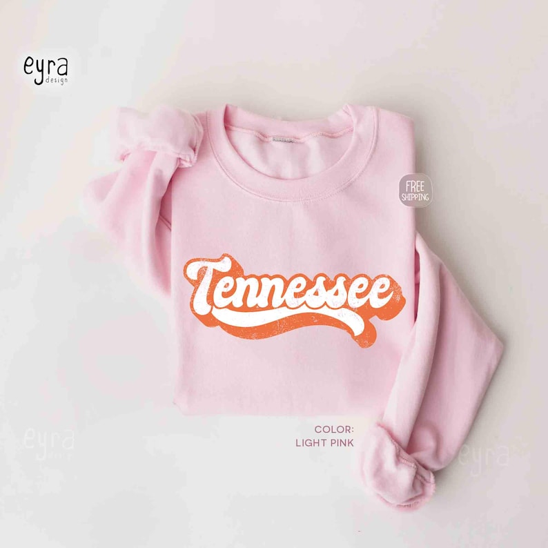 K&ouml;nnte beinhalten: Ein hellrosa Rundhals-Sweatshirt mit dem Wort Tennessee, das in einer kr&auml;ftigen, orangefarbenen Retro-Schrift mit wei&szlig;er Umrandung aufgedruckt ist. Das Kleidungsst&uuml;ck hat lange &Auml;rmel und einen gerippten Kragen mit einem klaren Design.