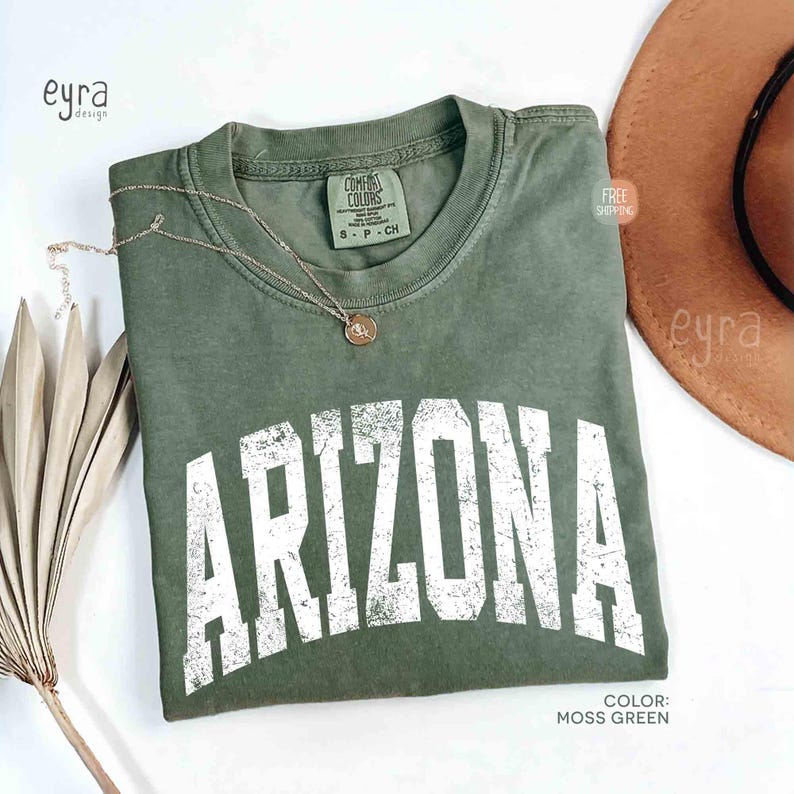 May include: A moss green short-sleeve t-shirt with a crew neckline featuring the word ARIZONA printed in large, distressed white capital letters across the chest. The shirt has a Comfort Colors brand label inside the collar and a thin gold necklace.
