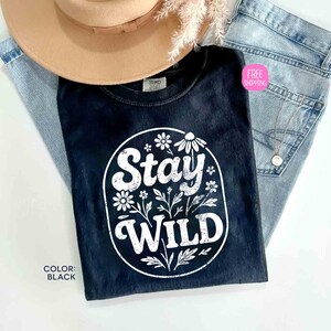 May include: Black t-shirt with the words "Stay Wild" in a white floral design. The shirt is paired with light-wash denim jeans and a tan hat. The shirt's color is black.