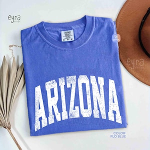 May include: A periwinkle blue short-sleeve t-shirt with a crew neckline. The front features the word ARIZONA printed in a large, distressed white collegiate-style font. The shirt has a Comfort Colors brand label at the inner neck area.