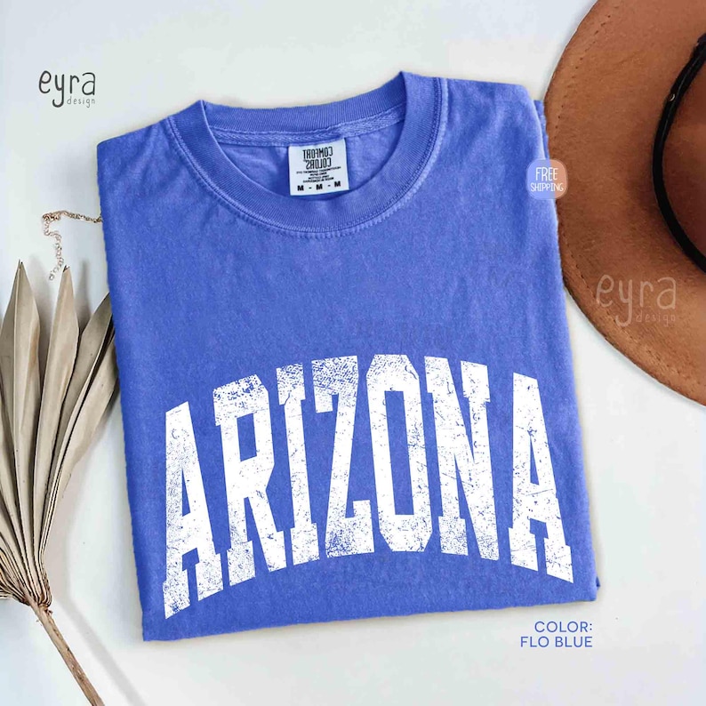 May include: A periwinkle blue short-sleeve t-shirt with a crew neckline. The front features the word ARIZONA printed in a large, distressed white collegiate-style font. The shirt has a Comfort Colors brand label at the inner neck area.