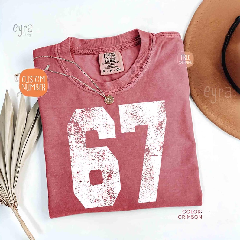 K&ouml;nnte beinhalten: Karmesinrotes T-Shirt mit der wei&szlig;en Zahl 67 im Used-Look. Das Shirt hat einen Rundhalsausschnitt und kurze &Auml;rmel. Eine silberne Halskette mit Anh&auml;nger liegt &uuml;ber dem Shirt. Das Etikett lautet "Comfort Colors".
