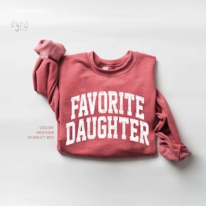 Op de afbeelding: Een heather scharlakenrood sweatshirt met lange mouwen en een ronde hals, voorzien van de tekst FAVORITE DAUGHTER in een verweerd wit lettertype in universiteitsstijl op de borst. Het kledingstuk heeft geribbelde boorden en een geribbelde halslijn.
