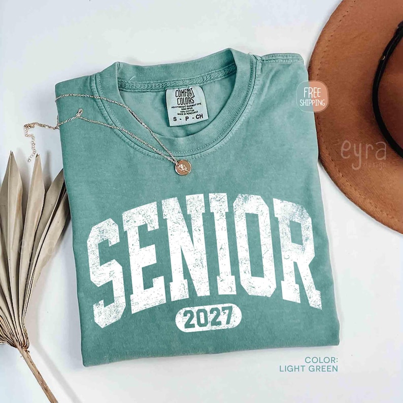 May include: Light green t-shirt with the word "SENIOR" in white block letters and the year "2027" below. A gold necklace with a pendant rests on the shirt. The shirt is labeled "Comfort Colors" and is size S-P-CH.