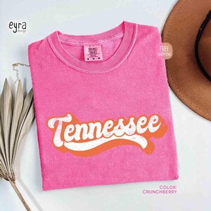 May include: A pink t-shirt with the word "Tennessee" in white script with an orange outline. The shirt has a solid colour and a vintage look. The colour is "Crunchberry".