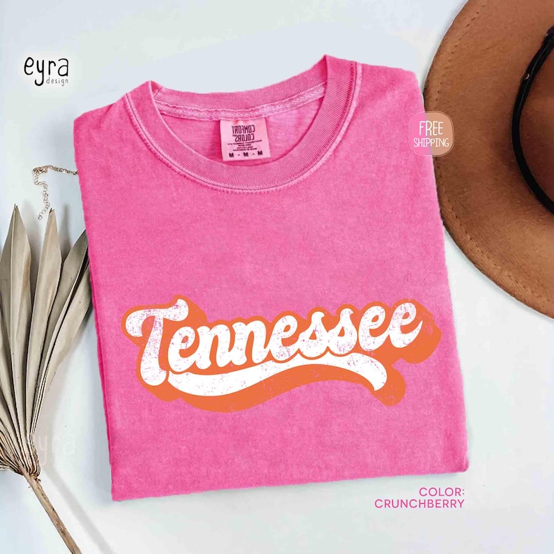 May include: A pink t-shirt with the word "Tennessee" in white script with an orange outline. The shirt has a solid colour and a vintage look. The colour is "Crunchberry".