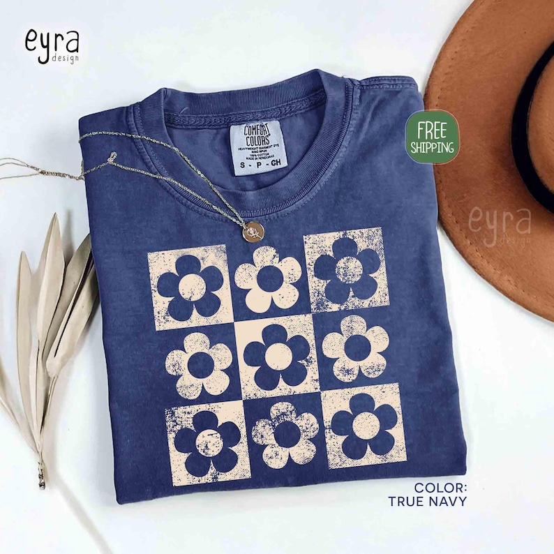 May include: A navy blue t-shirt with a retro floral design. The shirt features a grid of nine cream-colored flowers. A gold necklace rests on the shirt. The shirt is labeled "Comfort Colors" and the color is "True Navy".