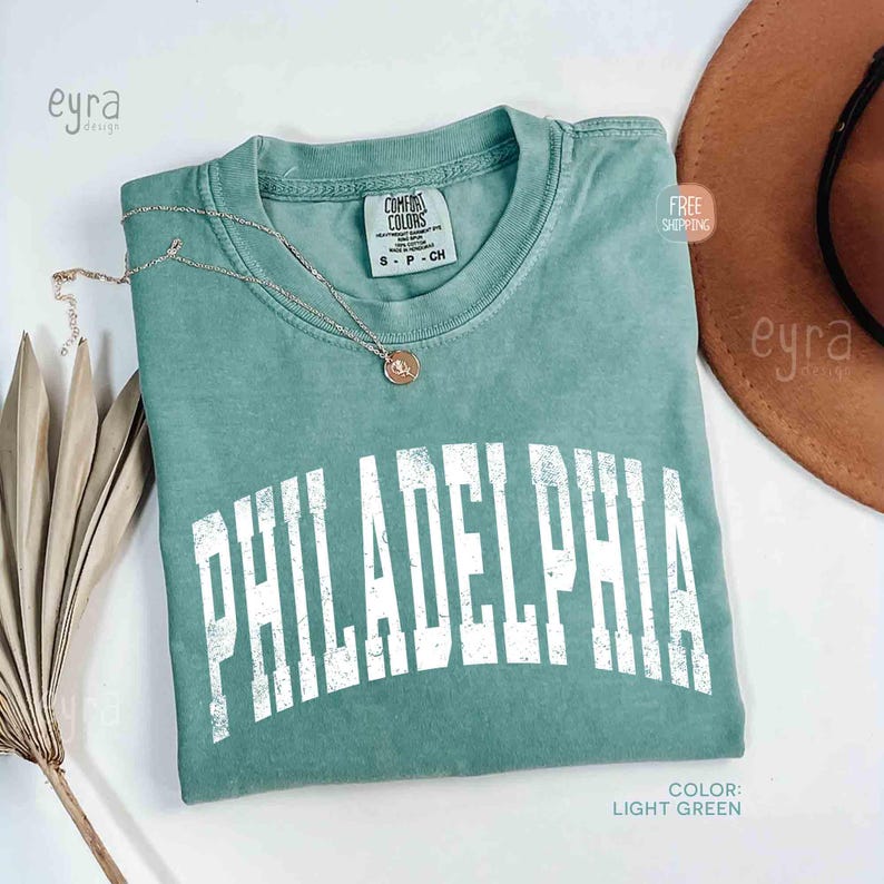 May include: A light green crew neck t-shirt featuring the word PHILADELPHIA printed in a large, distressed white collegiate font across the chest. The shirt has a Comfort Colors brand label at the inner neckline and a thin gold-toned necklace.