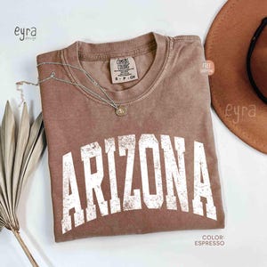 May include: A short-sleeved espresso brown t-shirt with a crew neckline and the word ARIZONA printed in large, distressed white block letters across the chest. The shirt features a Comfort Colors brand label on the inside collar.