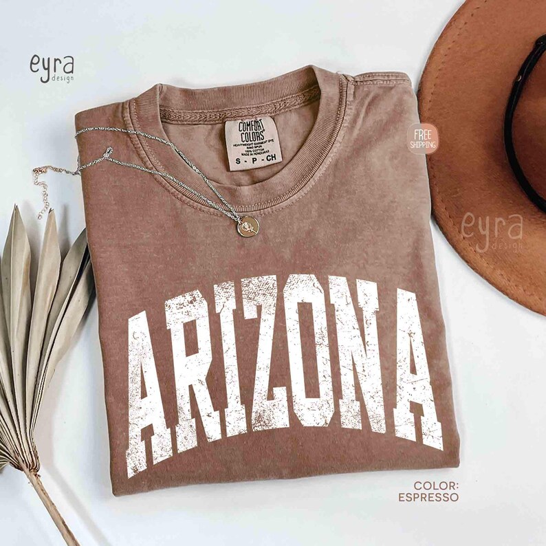 May include: A short-sleeved espresso brown t-shirt with a crew neckline and the word ARIZONA printed in large, distressed white block letters across the chest. The shirt features a Comfort Colors brand label on the inside collar.