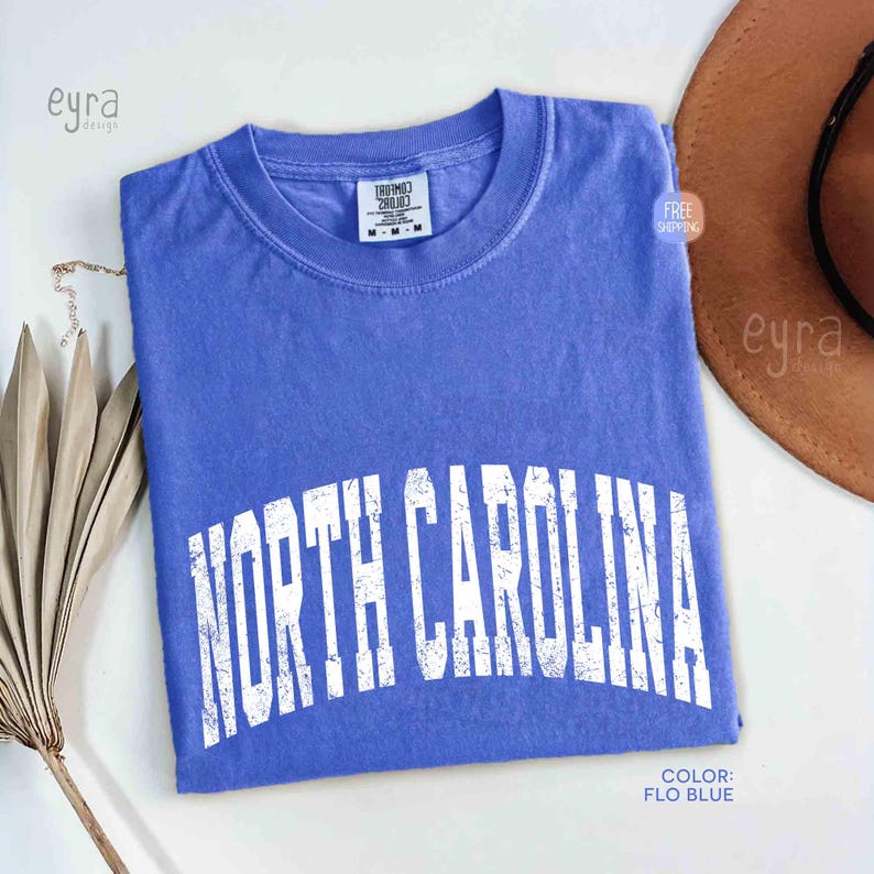 K&ouml;nnte beinhalten: Ein kurz&auml;rmeliges T-Shirt in der Farbe Kornblumenblau mit dem Schriftzug NORTH CAROLINA in einer verwaschenen, wei&szlig;en, bogenf&ouml;rmigen Schrift auf der Brust. Das Shirt hat einen Rundhalsausschnitt und ein Comfort Colors Etikett am Kragen.