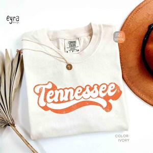 May include: Cream-coloured t-shirt featuring the word "Tennessee" in an orange retro font. A gold necklace with a pendant rests on the shirt. A brown hat is partially visible in the background. The shirt is labelled "Comfort Colors".
