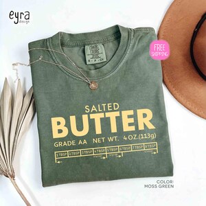 May include: Moss green t-shirt with the text "SALTED BUTTER" in yellow. The shirt includes a measurement guide and the text "GRADE AA NET WT. 4 OZ. (113g)". A gold necklace is draped on the shirt.
