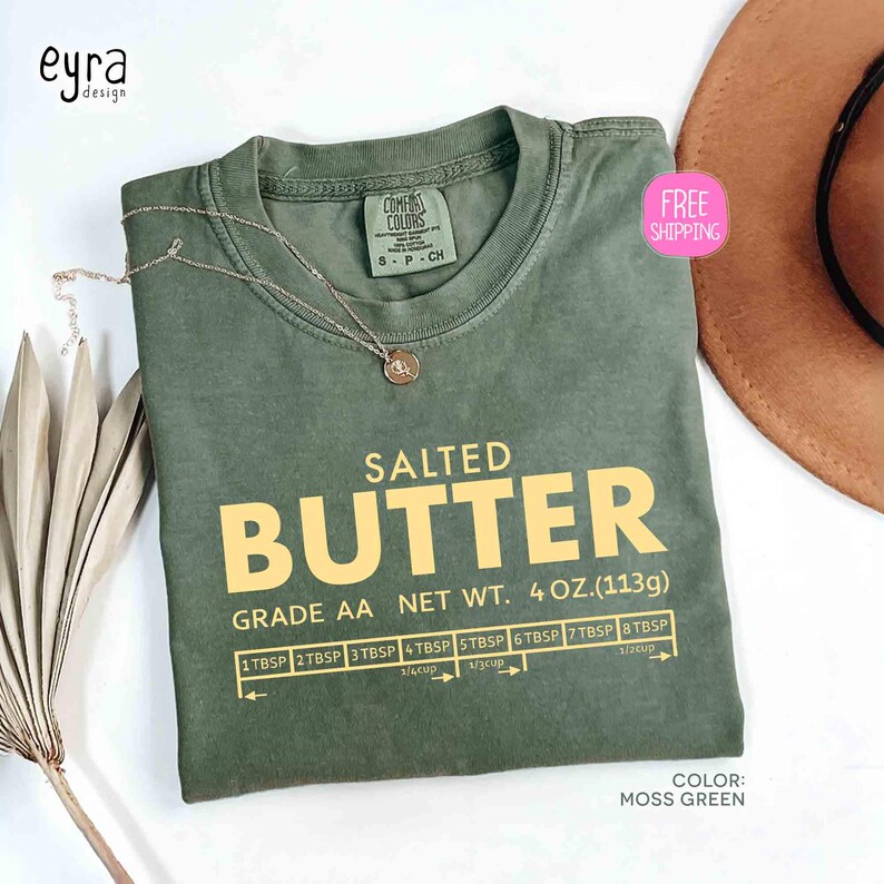 May include: Moss green t-shirt with the text "SALTED BUTTER" in yellow. The shirt includes a measurement guide and the text "GRADE AA NET WT. 4 OZ. (113g)". A gold necklace is draped on the shirt.