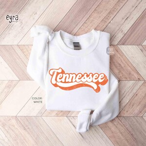 K&ouml;nnte beinhalten: Ein wei&szlig;es Langarm-Sweatshirt mit Rundhalsausschnitt und dem Wort Tennessee in einer fetten, orangefarbenen, kursiven Retro-Schrift auf der Brust. Das Kleidungsst&uuml;ck hat einen gerippten Kragen, B&uuml;ndchen und Saum sowie eine schlichte, wei&szlig;e Stoffoberfl&auml;che.