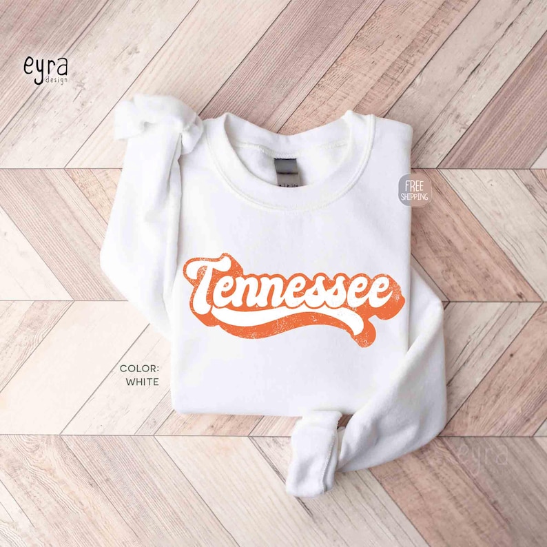 K&ouml;nnte beinhalten: Ein wei&szlig;es Langarm-Sweatshirt mit Rundhalsausschnitt und dem Wort Tennessee in einer fetten, orangefarbenen, kursiven Retro-Schrift auf der Brust. Das Kleidungsst&uuml;ck hat einen gerippten Kragen, B&uuml;ndchen und Saum sowie eine schlichte, wei&szlig;e Stoffoberfl&auml;che.