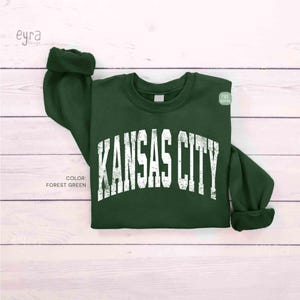 May include: A forest green sweatshirt featuring the text "KANSAS CITY" in distressed white lettering. The sweatshirt has a crew neck and cuffed sleeves. The colour is forest green.